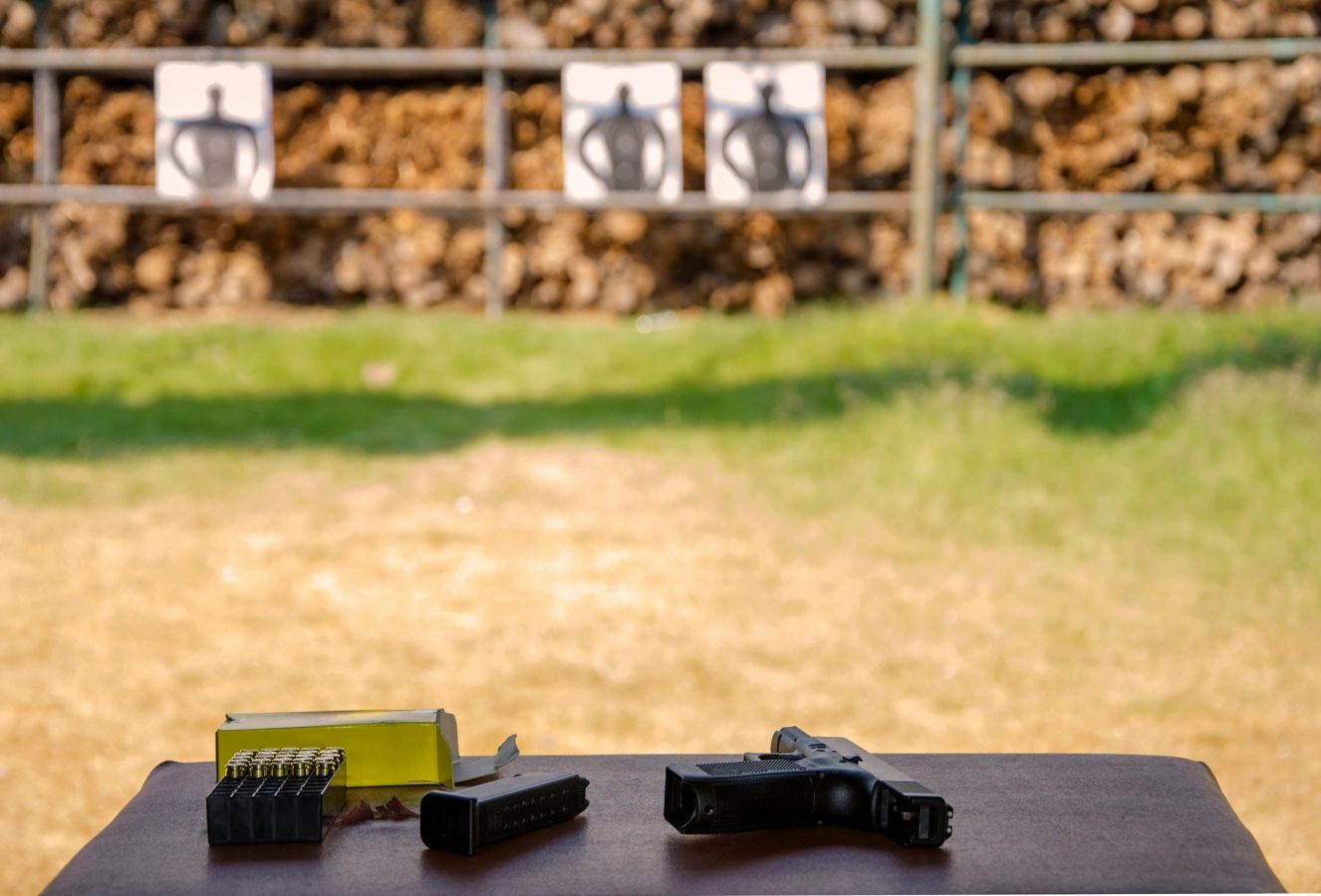Stock photo, illustrative. A pistol, a magazine, and a box of ammunition on a table, with silhouette targets behind them.
